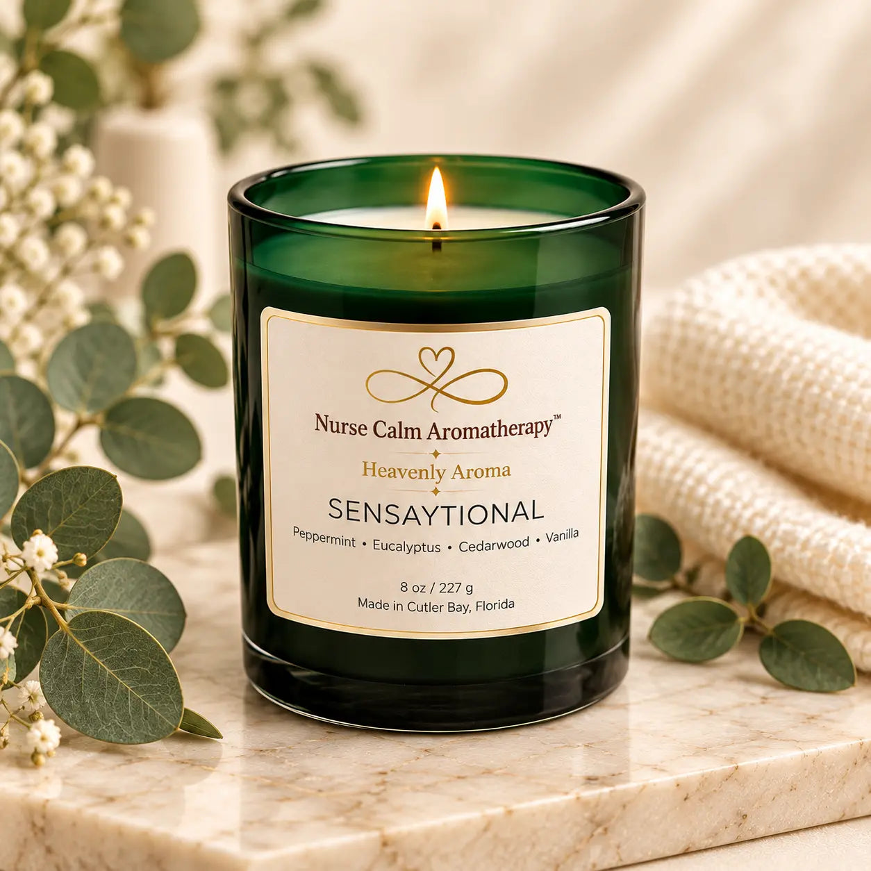 Lit Sensaytional 8 oz peppermint and eucalyptus soy candle by Nurse Calm Aromatherapy™, featuring a deep emerald-green glass jar with an ivory and gold label, displayed on a cream marble surface with eucalyptus greenery, delicate white flowers, and soft spa towels.
