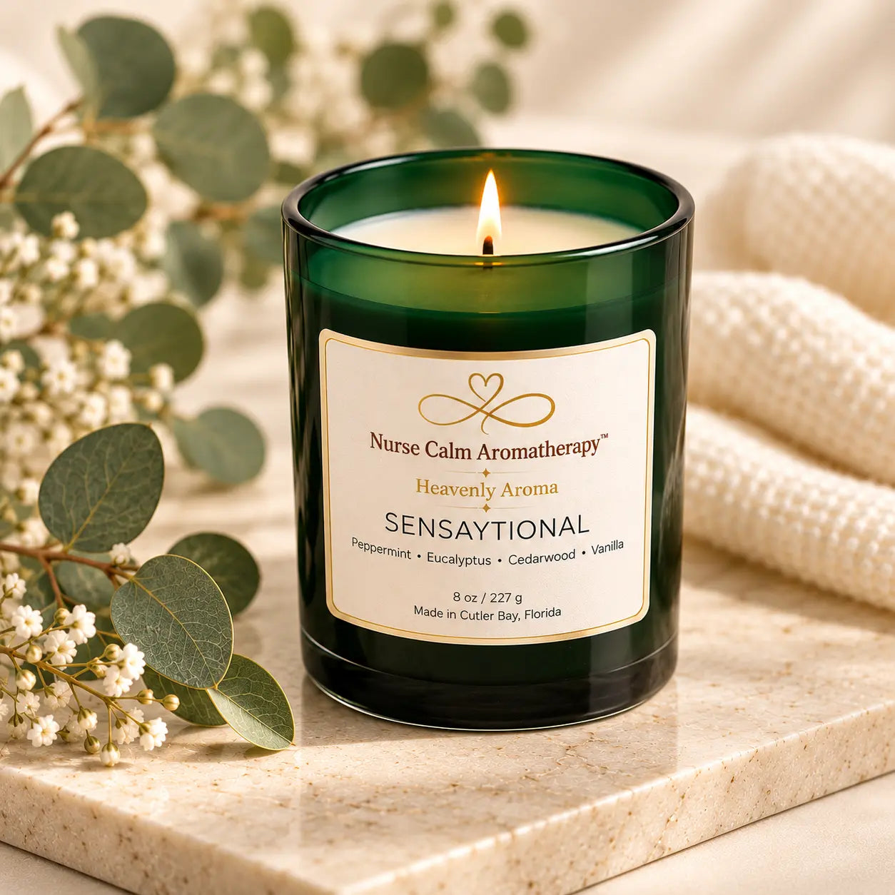 Angled view of the Sensaytional aromatherapy candle in a green glass jar, displayed on a cream marble surface with eucalyptus leaves and soft neutral accents.