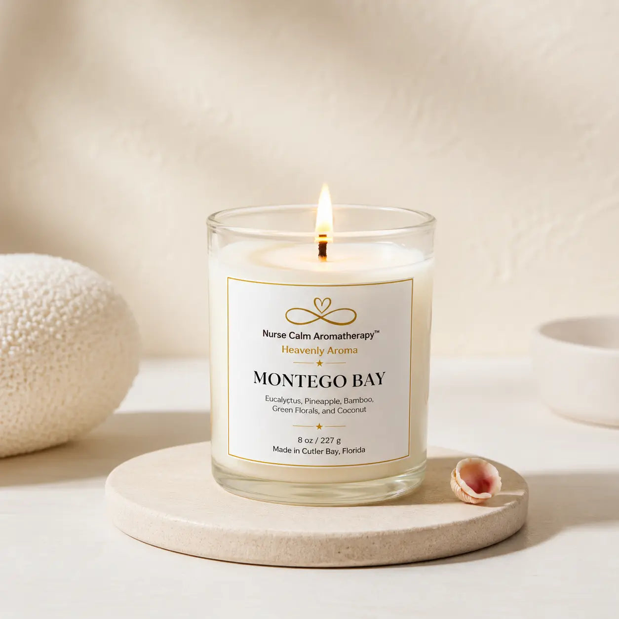 A lit candle in a clear glass jar sits on a round stone coaster, with a small seashell beside it and a softly textured neutral background. The label reads 'Montego Bay' and 'Nurse Calm Aromatherapy Heavenly Aromas.