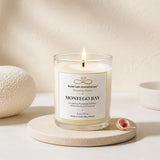 A lit candle in a clear glass jar sits on a round stone coaster, with a small seashell beside it and a softly textured neutral background. The label reads 'Montego Bay' and 'Nurse Calm Aromatherapy Heavenly Aromas.