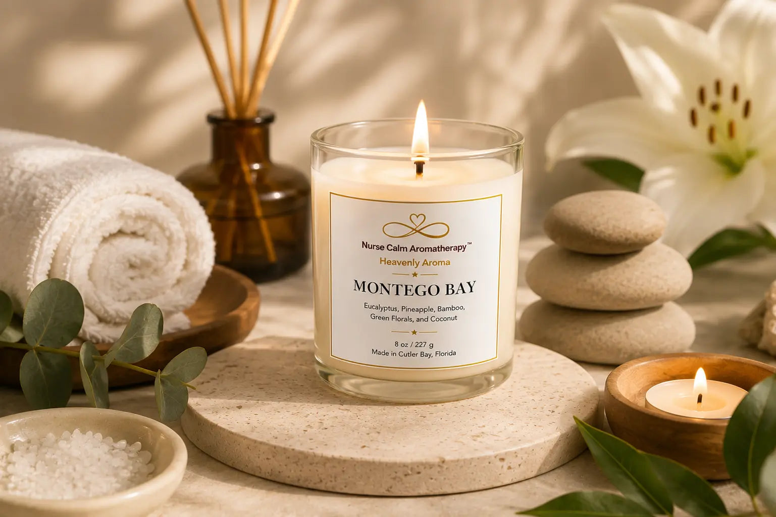 A lit glass jar candle labeled 'Montego Bay' by Nural Calm Aromatherapy, styled on a stone coaster with spa props including a rolled towel, diffuser, stacked stones, bath salts, and greenery.