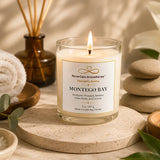 A lit glass jar candle labeled 'Montego Bay' by Nural Calm Aromatherapy, styled on a stone coaster with spa props including a rolled towel, diffuser, stacked stones, bath salts, and greenery.