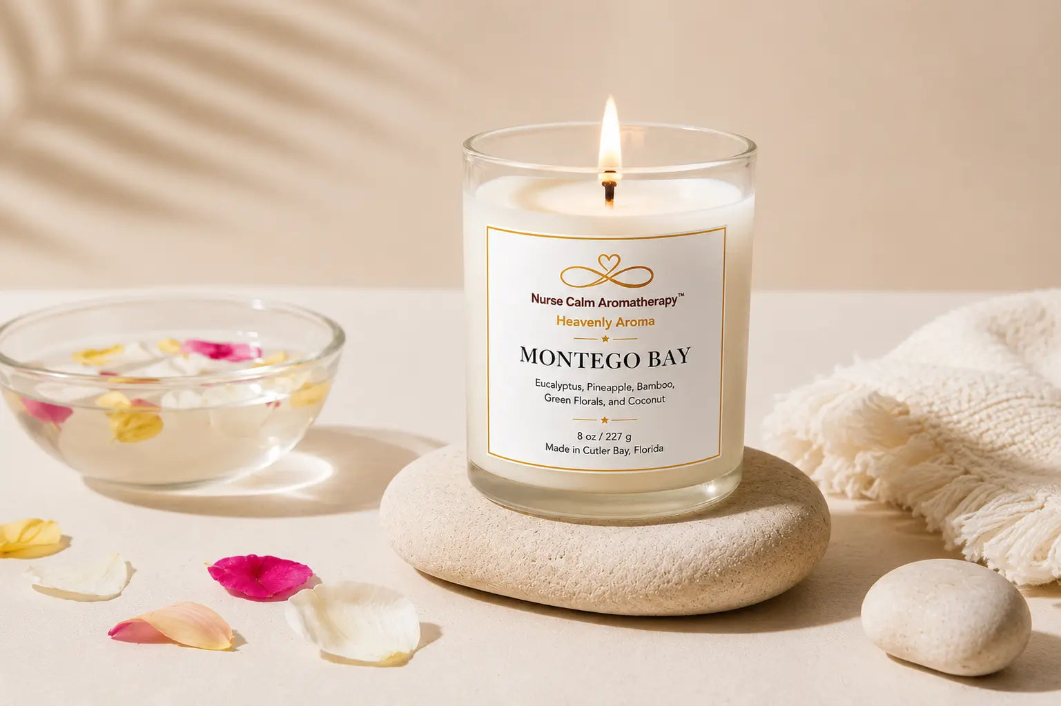 A lit glass jar candle labeled 'Montego Bay' by Nurse Calm Aromatherapy sits on a round stone coaster, styled with a bowl of flower petals, scattered petals, and a soft cream towel on a warm beige surface.