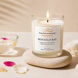 A lit glass jar candle labeled 'Montego Bay' by Nurse Calm Aromatherapy sits on a round stone coaster, styled with a bowl of flower petals, scattered petals, and a soft cream towel on a warm beige surface.