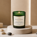 Sensaytional soy candle by Nurse Calm Aromatherapy™ displayed on a neutral cream pedestal with soft spa-inspired décor. Green glass candle with gold-trimmed label and lit flame featuring peppermint, eucalyptus, cedarwood, and vanilla fragrance notes.
