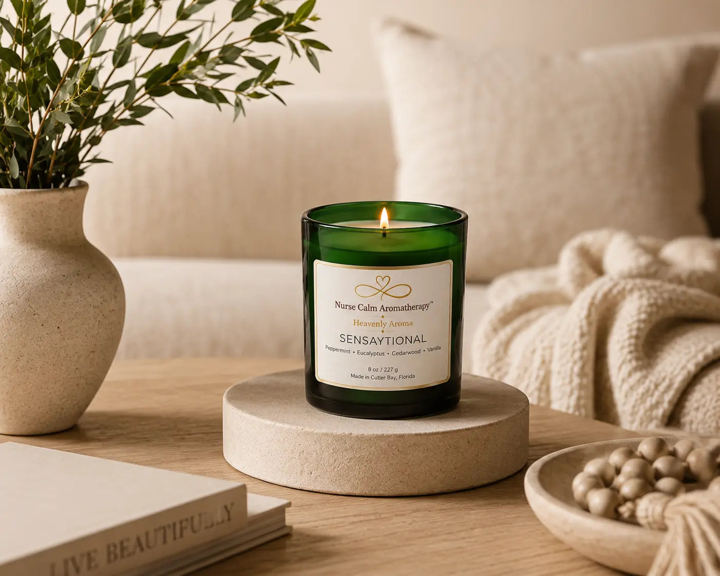 Sensaytional soy candle by Nurse Calm Aromatherapy™ displayed in a cozy neutral-toned living space with soft blankets, natural textures, and warm ambient lighting. Green glass candle with lit flame featuring peppermint, eucalyptus, cedarwood, and vanilla fragrance notes.
