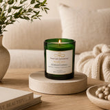Sensaytional soy candle by Nurse Calm Aromatherapy™ displayed in a cozy neutral-toned living space with soft blankets, natural textures, and warm ambient lighting. Green glass candle with lit flame featuring peppermint, eucalyptus, cedarwood, and vanilla fragrance notes.