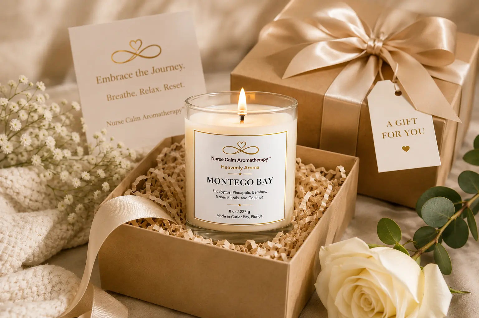 A lit jar candle labeled 'Montego Bay' by Nurse Cal Aromatherapy sits in a kraft gift box with shredded paper filler, alongside a wrapped gift box, a card, and white flowers on a soft beige background.