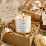 A lit jar candle labeled 'Montego Bay' by Nurse Cal Aromatherapy sits in a kraft gift box with shredded paper filler, alongside a wrapped gift box, a card, and white flowers on a soft beige background.