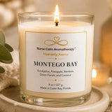 A lit candle in a clear glass jar with a white label reading 'Nurse Calm Aromatherapy' and 'Montego Bay,' styled on a wooden tray with soft warm lighting and eucalyptus leaves around it.